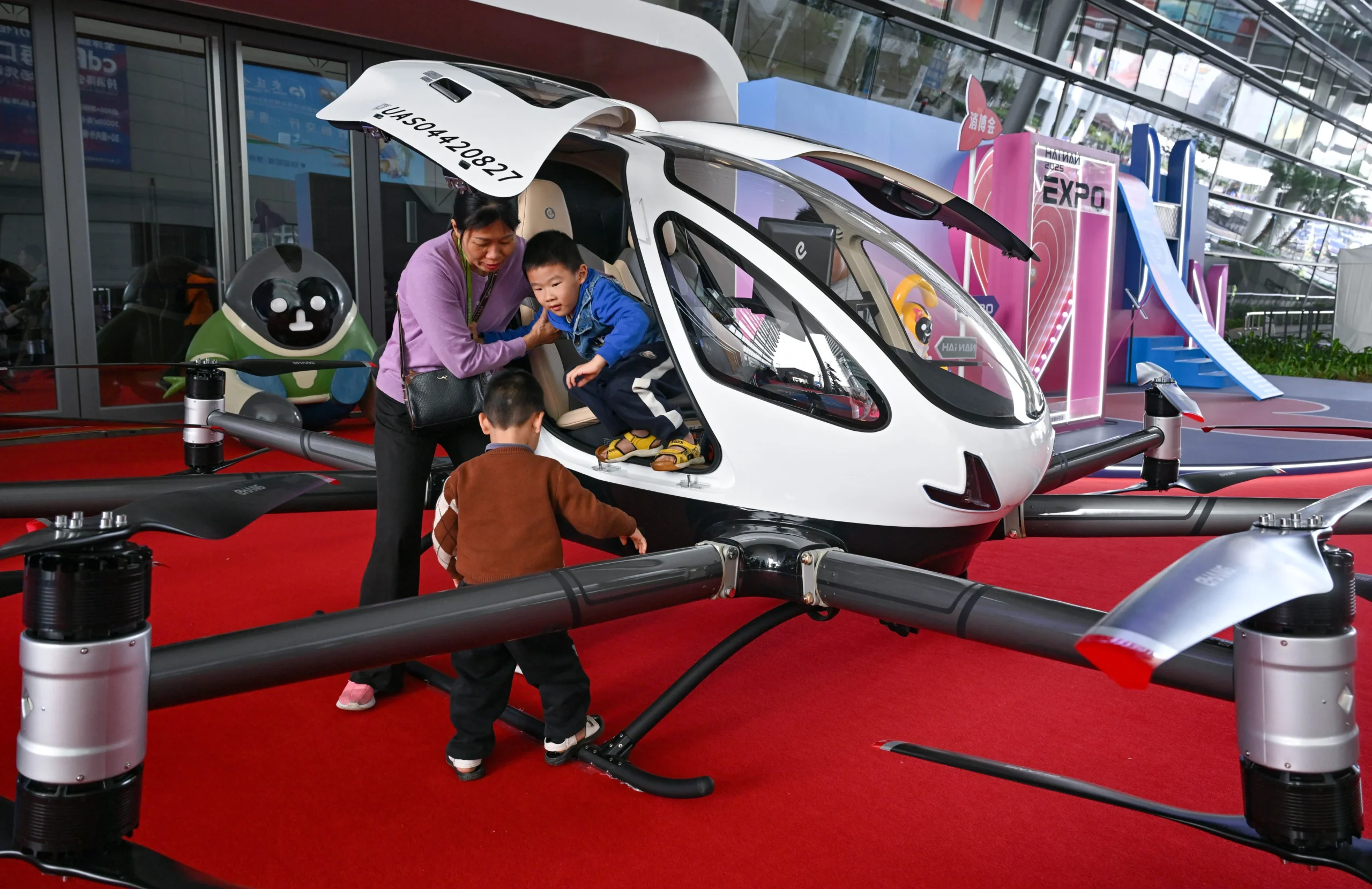 A woman helps a child exit a white autonomous passenger drone on display at a tech expo, with another child nearby.