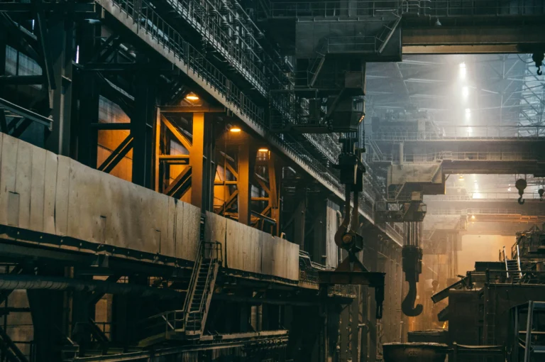 Interior of an industrial factory with metal structures, cranes, and dim lighting.